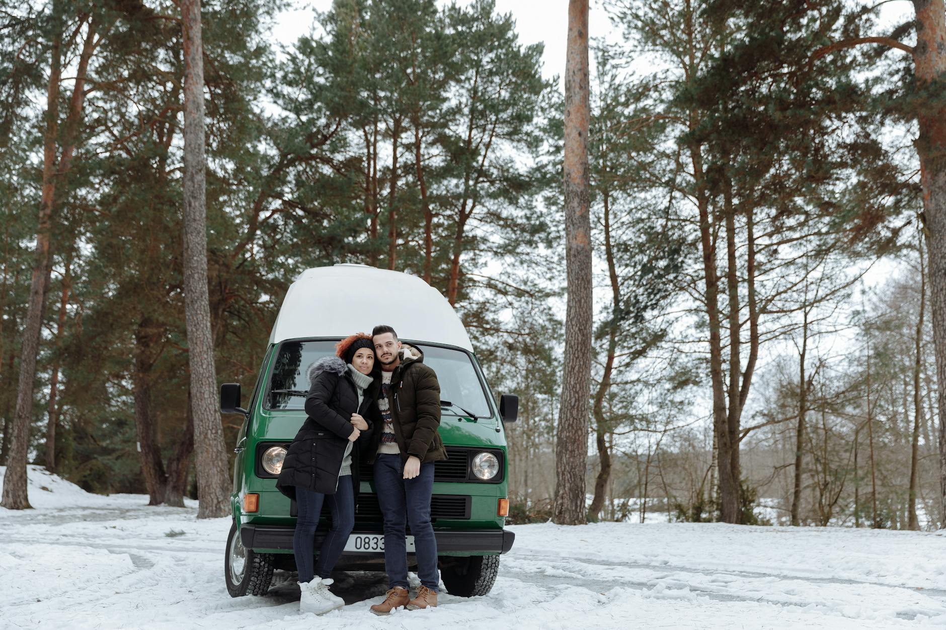 Couple in winter clothing embracing by a green campervan in a snowy forest setting, reflecting a cozy outdoor adventure.